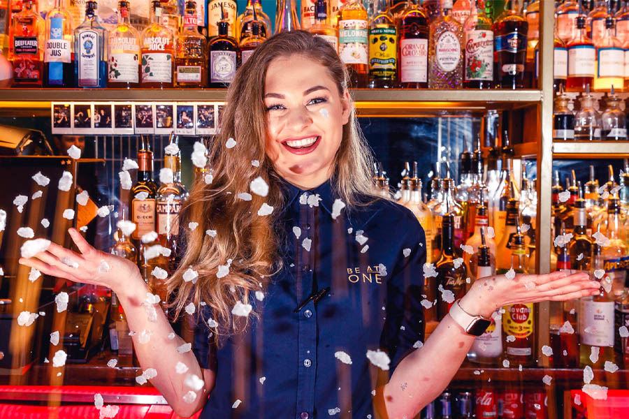 A woman standing in front of a bar with confetti falling from her hands.