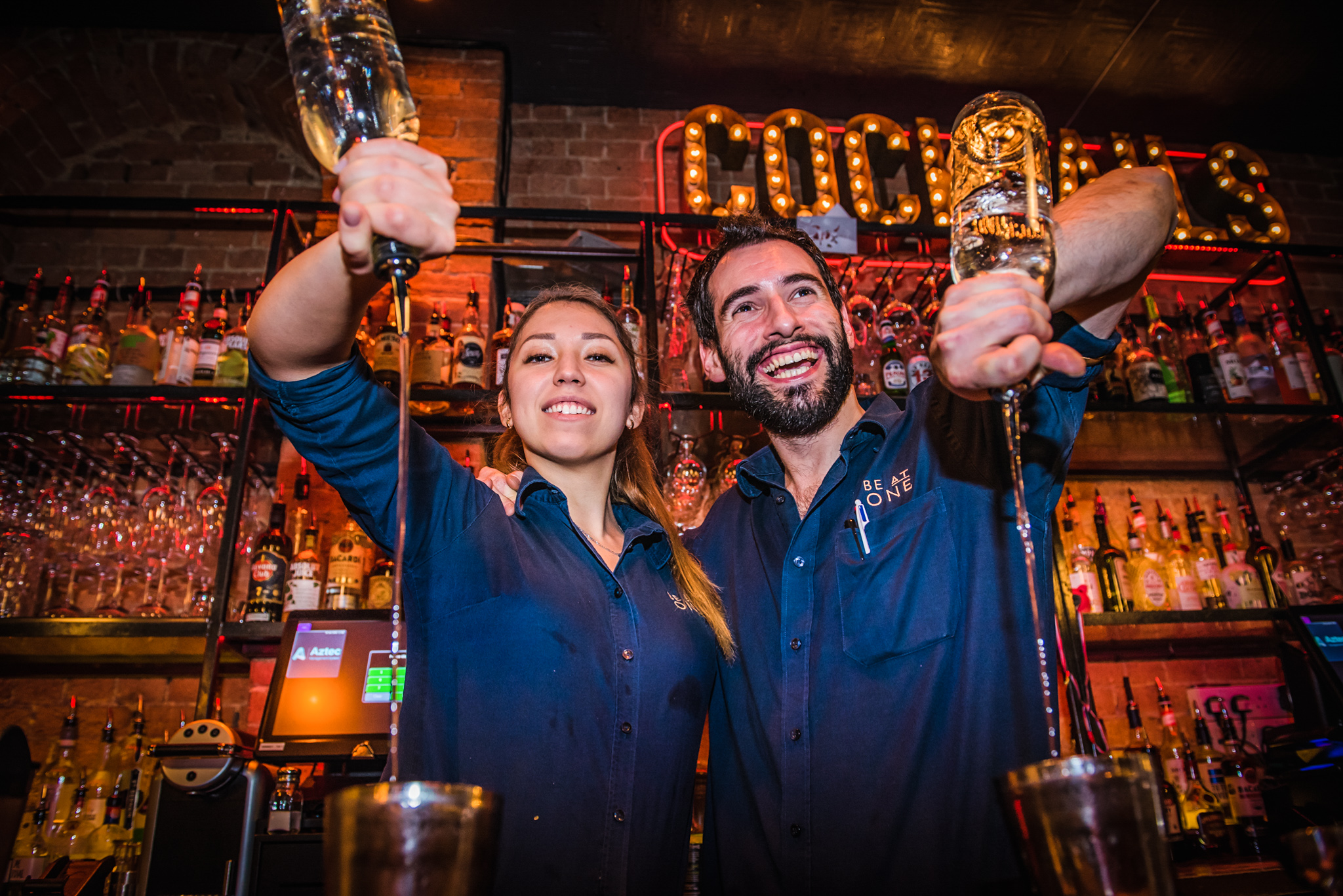 Two bartenders at a bar with a drink in their hands.